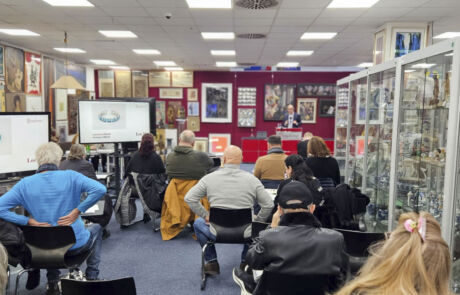Blick in ein Auktionshaus mit Menschen, die in Stuhlreihen auf einer Kunstauktion sitzen. Im Hintergrund Kunstwerke und Vitrinen mit Exponaten.