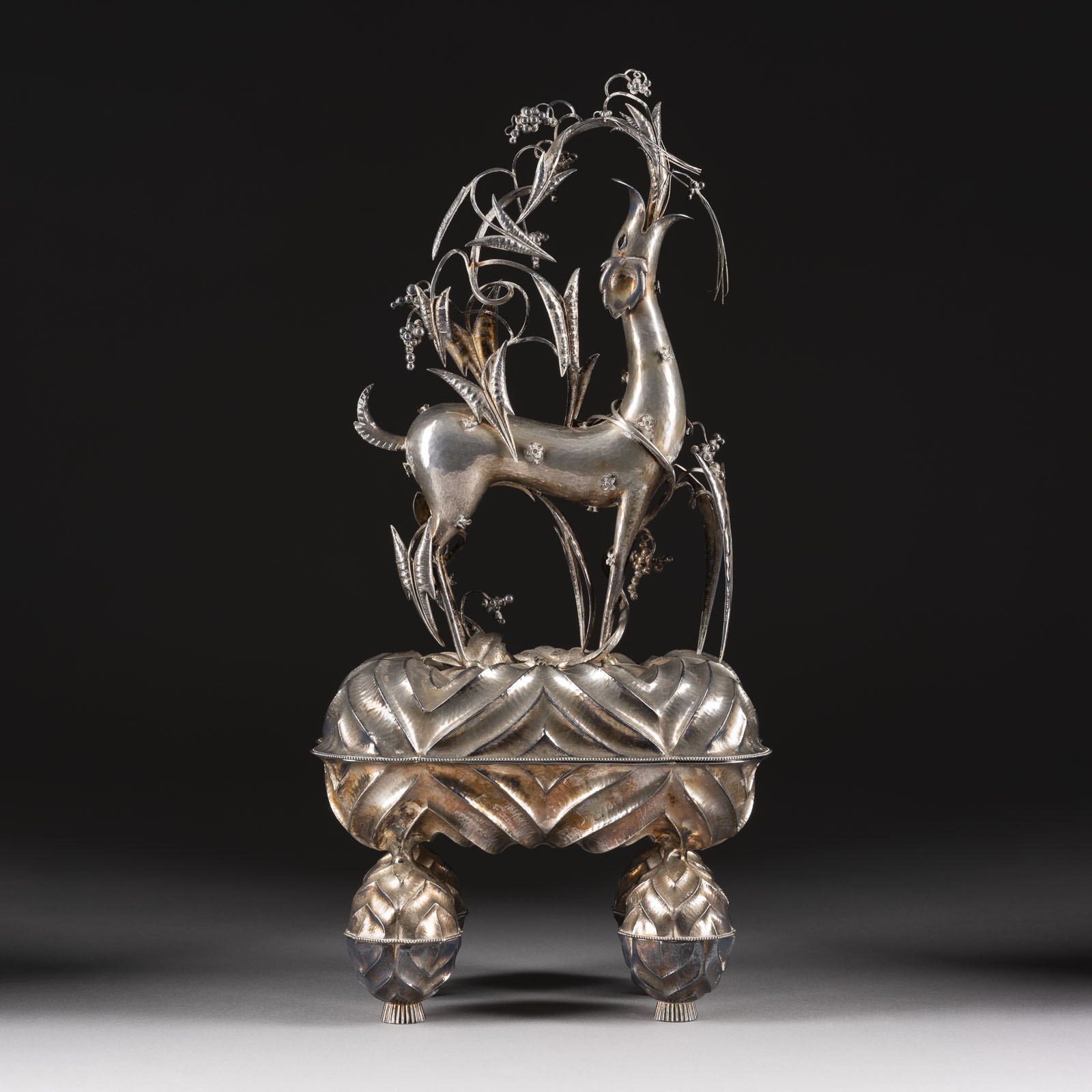 DAGOBERT PECHE HOCHBEDEUTENDE UND ÜBERAUS SELTENE SILBERDOSE ‚FABELTIER‘ DER WIENER WERKSTÄTTEN, 1920 Silberne Skulptur eines springenden Fabeltiers auf einem reich verzierten Podest, umgeben von filigranen Pflanzen und Blumen, vor neutralem Hintergrund.